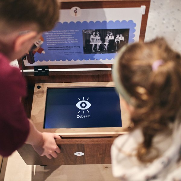 Dwoje dzieci z góry przygląda się interaktywnej drewnianej skrzyni w muzeum. Na wbudowanym ekranie świeci ikona oka z napisem „Zobacz”. Powyżej umieszczono tabliczkę informacyjną ze zdjęciem i tekstem.