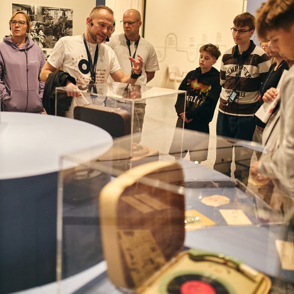Kurator wystawy opowiada grupie zwiedzających o eksponatach w muzeum. Na pierwszym planie widać przeszkloną gablotę, w której znajduje się dawny gramofon walizkowy z płytą winylową.