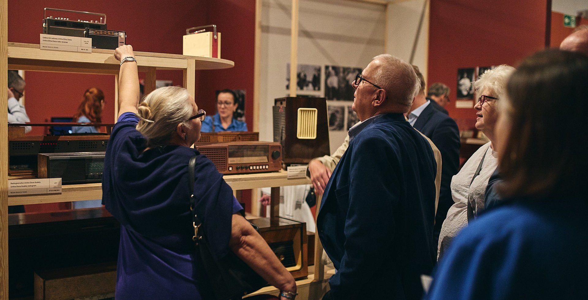 Zdjęcie przedstawia grupę starszych osób zwiedzających wystawę dawnych odbiorników radiowych w muzeum. Kobieta po lewej unosi rękę, wskazując na jedno z czarnych, prostokątnych seryjnych radiów stojących na drewnianym regale w sali o bordowych ścianach.