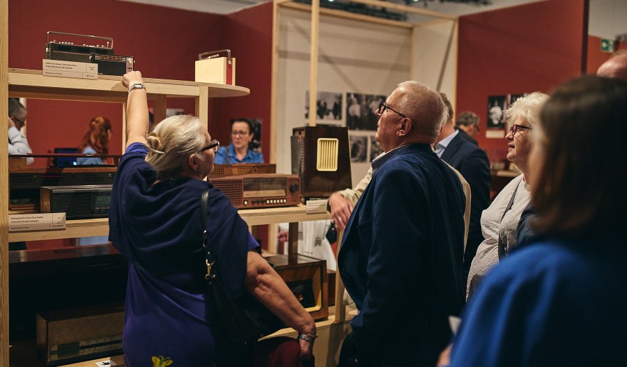Zdjęcie przedstawia grupę starszych osób zwiedzających wystawę dawnych odbiorników radiowych w muzeum. Kobieta po lewej unosi rękę, wskazując na jedno z czarnych, prostokątnych seryjnych radiów stojących na drewnianym regale w sali o bordowych ścianach.