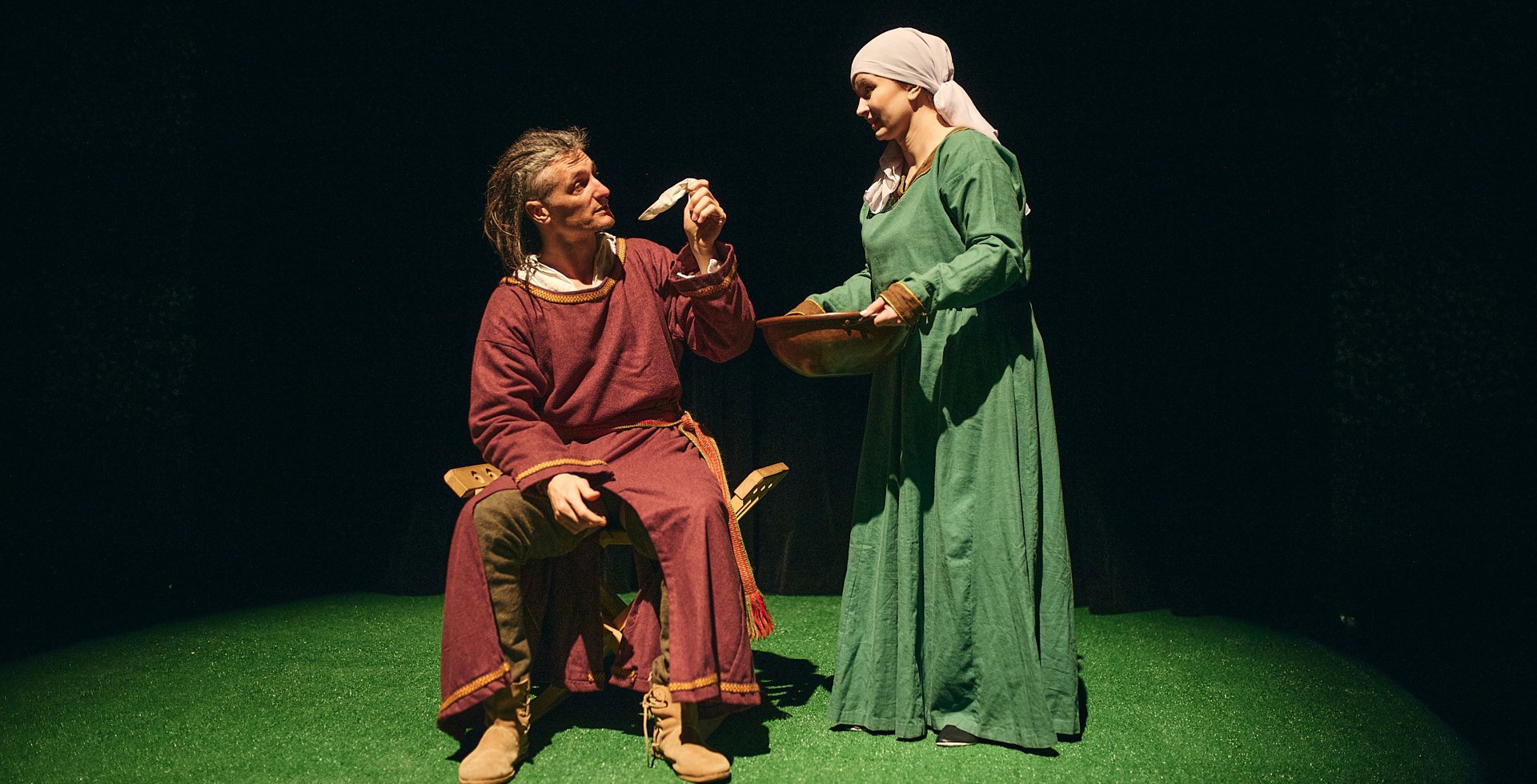 Scena teatralna na zielonej wykładzinie. Mężczyzna w bordowej szacie siedzi na stołku i trzyma w dłoni mały przedmiot. Obok niego stoi kobieta w zielonej sukni i białej chustce na głowie, trzymająca w dłoniach drewnianą misę. Tło jest czarne.