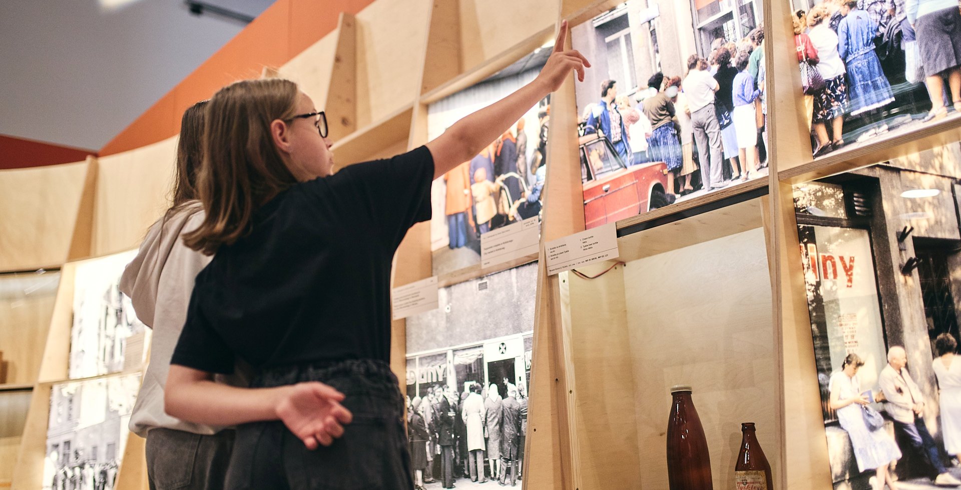 Dwie młode osoby oglądają wystawę historyczną w muzeum. Dziewczyna w okularach i czarnej koszulce unosi rękę, wskazując palcem na duże, archiwalne zdjęcie przedstawiające kolejkę ludzi przed sklepem mięsnym w czasach PRL-u.