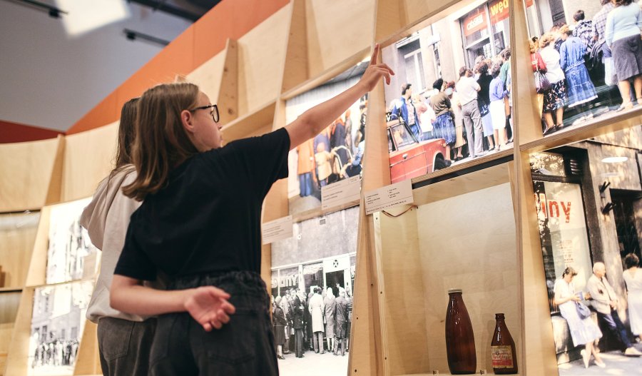 Dwie młode osoby oglądają wystawę historyczną w muzeum. Dziewczyna w okularach i czarnej koszulce unosi rękę, wskazując palcem na duże, archiwalne zdjęcie przedstawiające kolejkę ludzi przed sklepem mięsnym w czasach PRL-u.