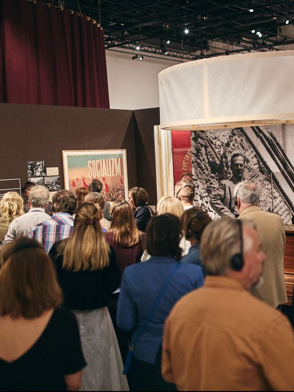 Tłum zwiedzających z audioprzewodnikami ogląda wystawę historyczną w muzeum. W tle widać brązowe ściany z plakatami z epoki socjalizmu oraz dużą, okrągłą instalację wiszącą pod sufitem. Ludzie stoją tyłem i bokiem do obiektywu.