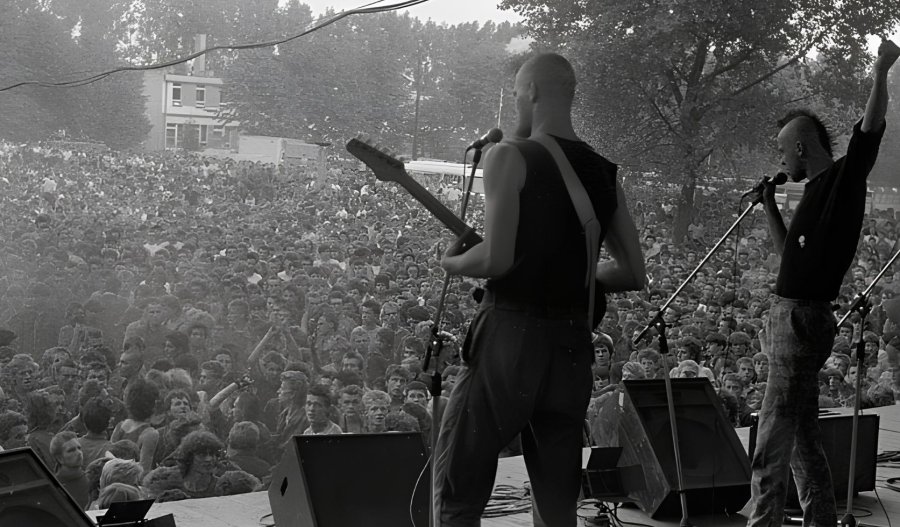 Czarno-białe zdjęcie z koncertu punkowego widziane zza pleców muzyków na scenie. Wokalista z irokezem śpiewa do mikrofonu z uniesioną pięścią, a obok stoi gitarzysta. Przed sceną bawi się ogromny, gęsty tłum publiczności.