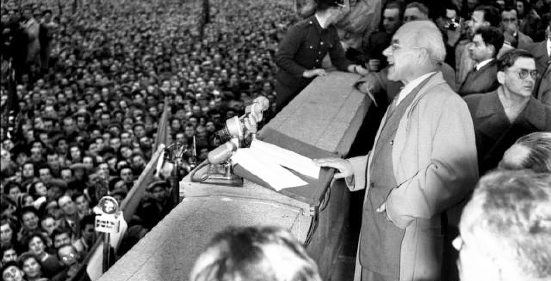 Władysław Gomułka w okularach i jasnym płaszczu przemawia z trybuny na warszawskim Placu Defilad 24 października 1956 roku. Przed nim stoją mikrofony, a w tle widać olbrzymi, gęsty tłum zgromadzonych ludzi, uczestniczących w wiecu Października '56.