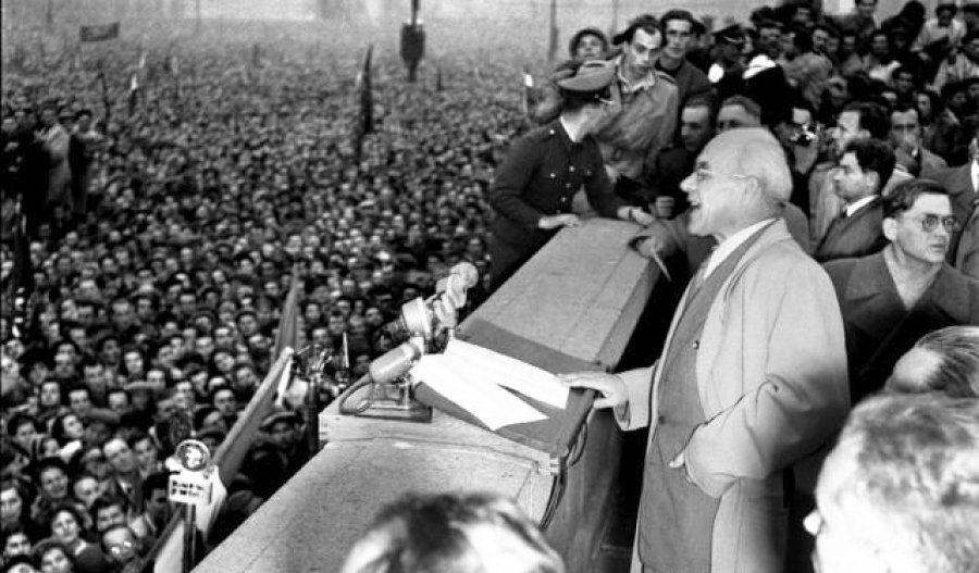 Władysław Gomułka w okularach i jasnym płaszczu przemawia z trybuny na warszawskim Placu Defilad 24 października 1956 roku. Przed nim stoją mikrofony, a w tle widać olbrzymi, gęsty tłum zgromadzonych ludzi, uczestniczących w wiecu Października '56.