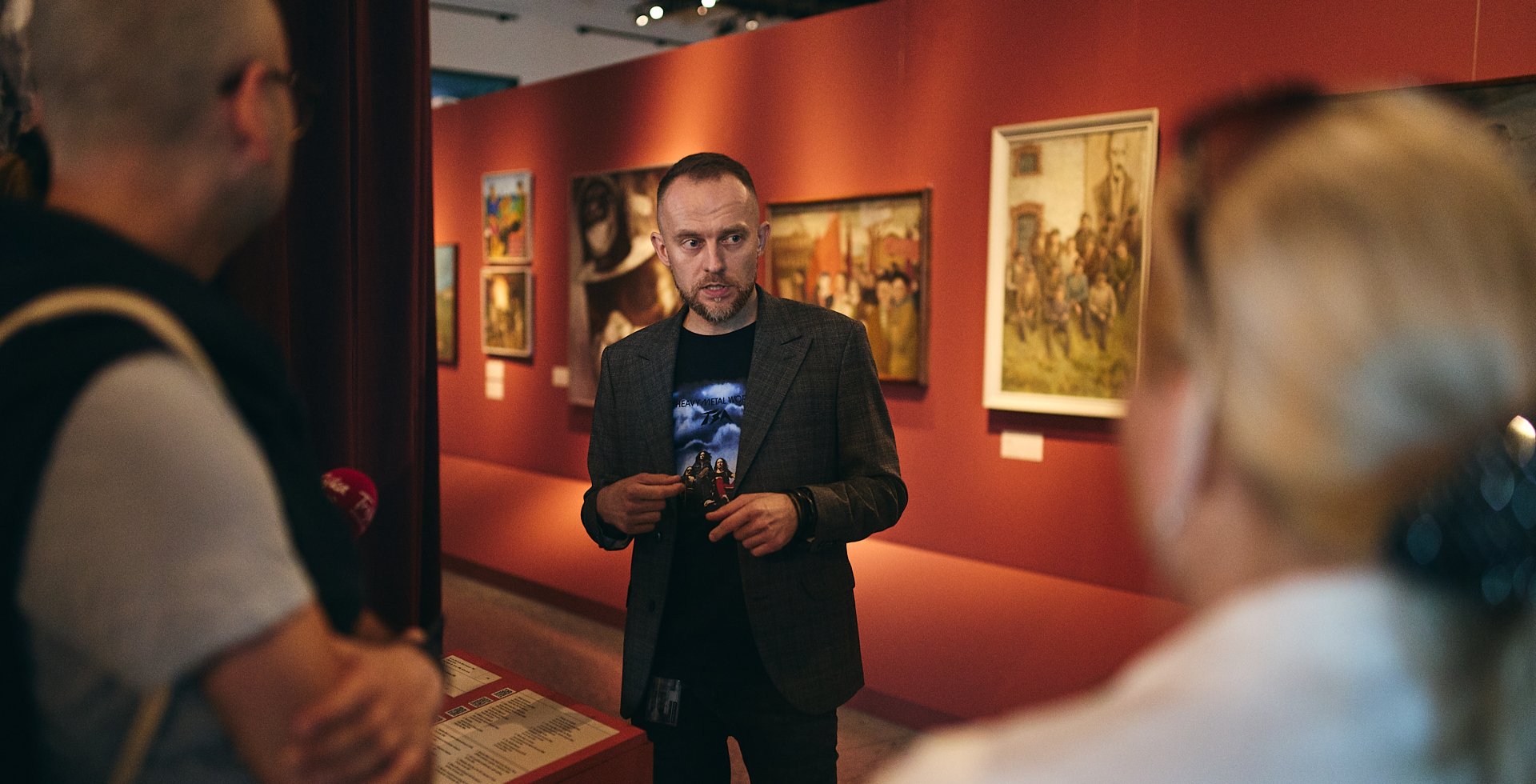 Mężczyzna w szarym garniturze i ciemnym t-shircie z nadrukiem przemawia do grupy osób w galerii sztuki. W tle widać czerwone ściany, na których wiszą obrazy w ramach. Kadrowane zza ramion słuchaczy, światło jest ciepłe i klimatyczne.