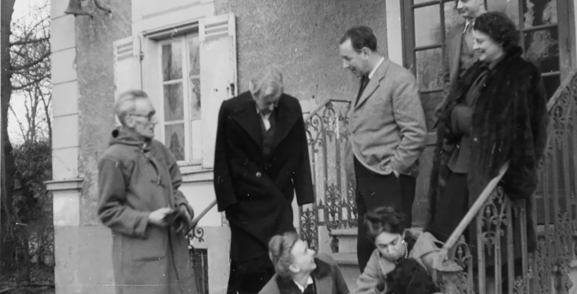 Czarno-białe archiwalne zdjęcie grupy osób na schodach willi w Maisons-Laffitte. Kobiety i mężczyźni w zimowych płaszczach rozmawiają i uśmiechają się do siebie, jedna z kobiet przytula psa. W tle fasada domu z okiennicami i kutą balustradą.