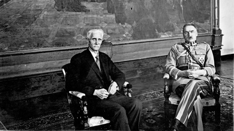 Na fotelach, na tle wielkiego obrazu siedzą Daszyński i Piłsudski. Daszyński jest w czarnym garniturze, a Piłsudski w mundurze wojskowym.