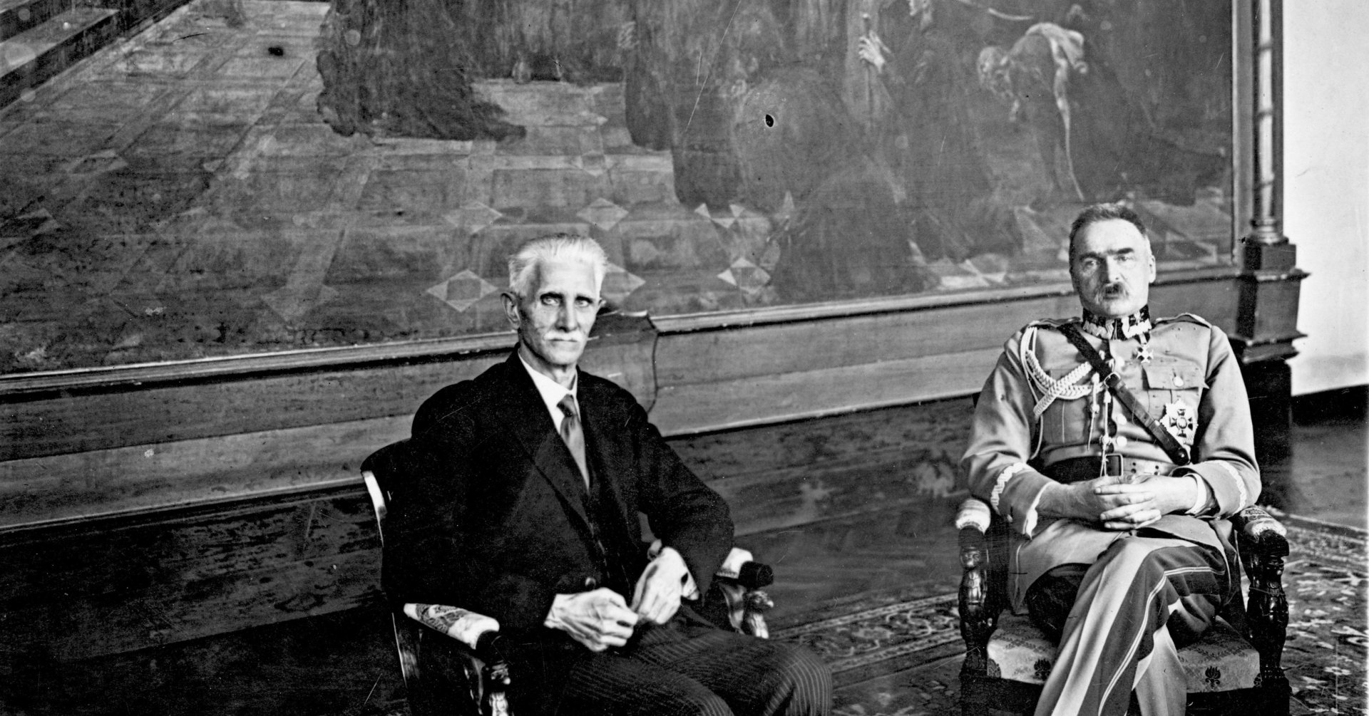Na fotelach, na tle wielkiego obrazu siedzą Daszyński i Piłsudski. Daszyński jest w czarnym garniturze, a Piłsudski w mundurze wojskowym.