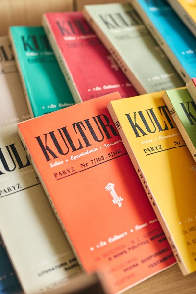Kolorowe zeszyty paryskiej „Kultury” ułożone na drewnianym stole, na pierwszym planie fragment wydruku ze starą reklamą.