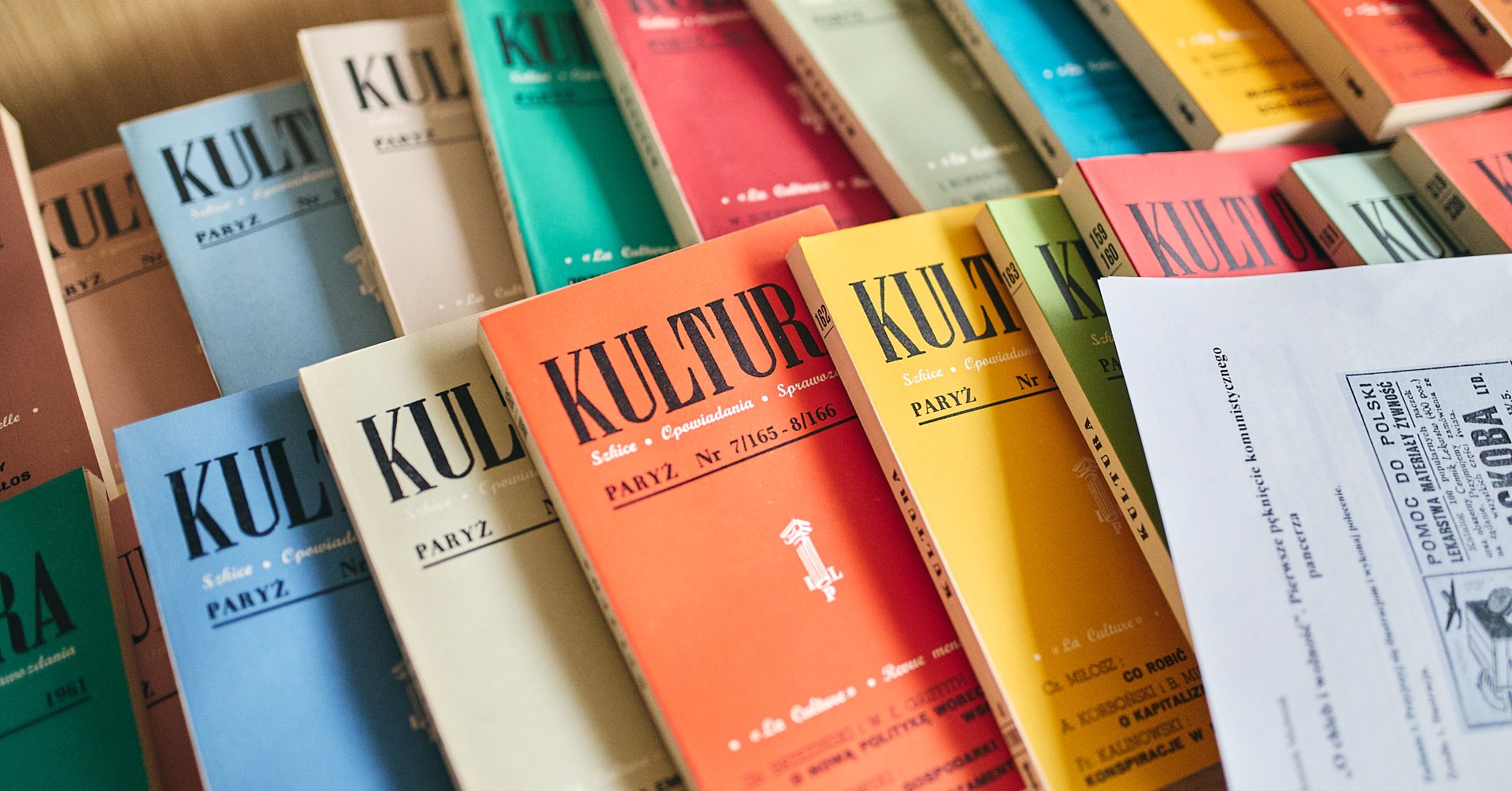Kolorowe zeszyty paryskiej „Kultury” ułożone na drewnianym stole, na pierwszym planie fragment wydruku ze starą reklamą.