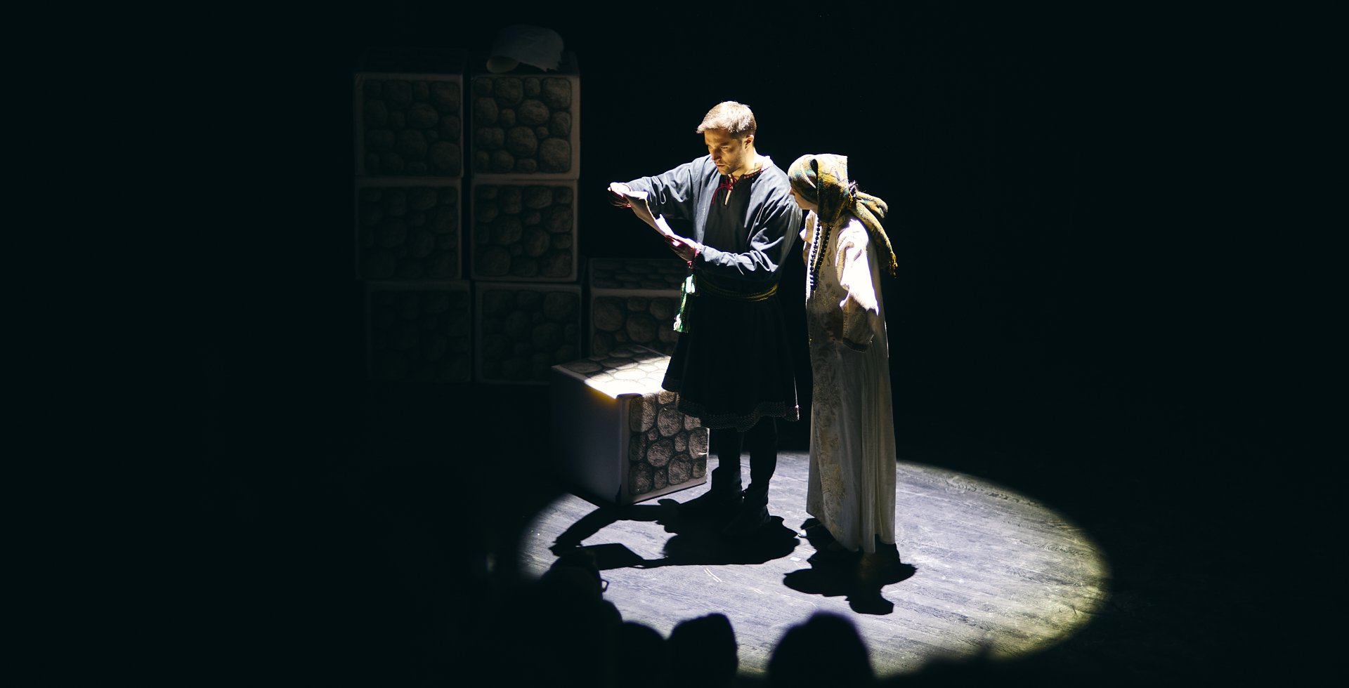 Scena teatralna na zielonej wykładzinie. Mężczyzna w bordowej szacie siedzi na stołku i trzyma w dłoni mały przedmiot. Obok niego stoi kobieta w zielonej sukni i białej chustce na głowie, trzymająca w dłoniach drewnianą misę. Tło jest czarne.