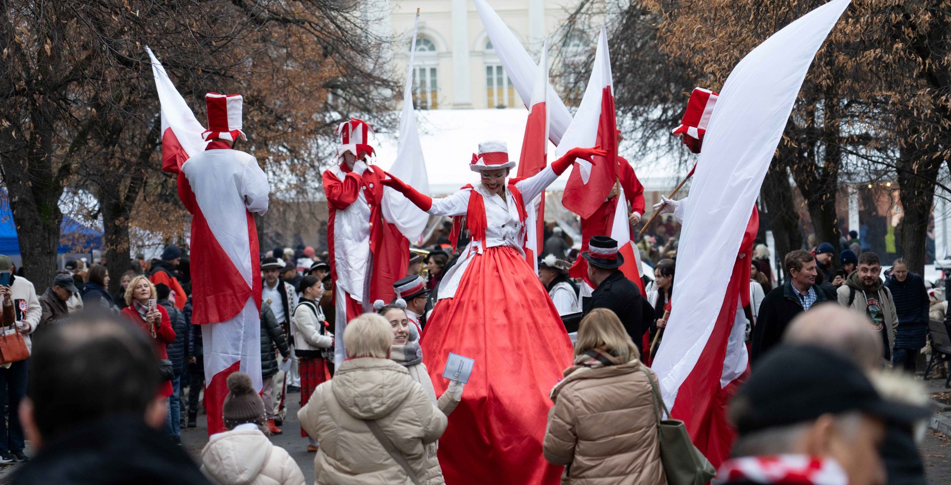 Uczestnicy parady z okazji Święta Niepodległości na Krakowskim Przedmieściu w Warszawie. Animatorzy na szczudłach w biało-czerwonych strojach tańczą i machają flagami Polski. Wokół gromadzą się przechodnie, w tle widać historyczny budynek.