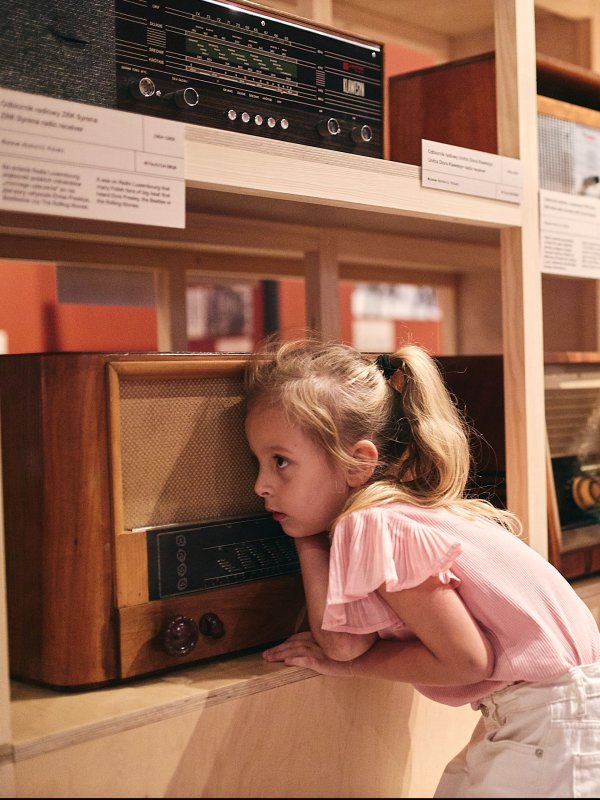Trzy małe dziewczynki z zaciekawieniem nasłuchują dźwięków ze starych, drewnianych odbiorników radiowych. Radia stoją na jasnym, drewnianym regale w muzeum. Dzieci przykładają uszy blisko głośników, a jedna z nich trzyma pluszową zabawkę.