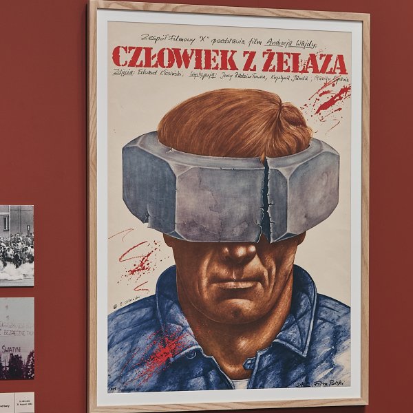 Plakat filmu Andrzeja Wajdy „Człowiek z żelaza” w drewnianej ramie na bordowej ścianie w muzeum. Przedstawia mężczyznę z pękniętą, metalową śrubą na oczach. Obok wiszą inne dokumenty i fragment niebieskiego plakatu z godłem Polski.