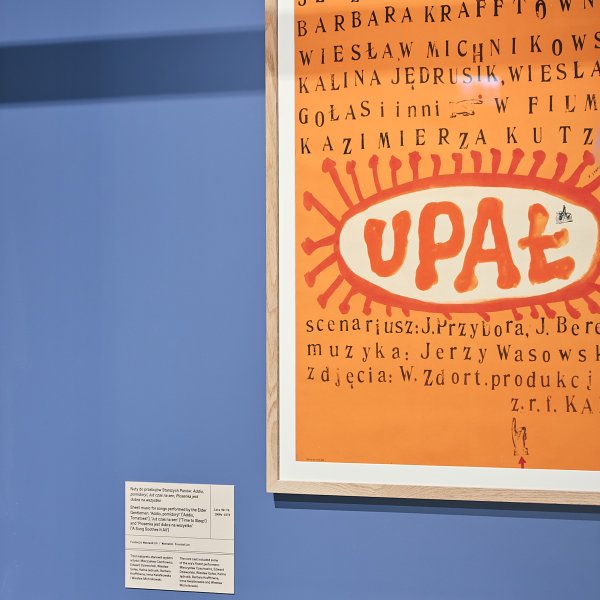 Plakat filmu „Upał” w drewnianej ramie na jasnoniebieskiej ścianie muzealnej. Widnieje na nim duże, pomarańczowe słońce z napisem oraz nazwiska twórców, m.in. Jeremiego Przybory. Poniżej znajduje się mała tabliczka informacyjna i szklana gablota.