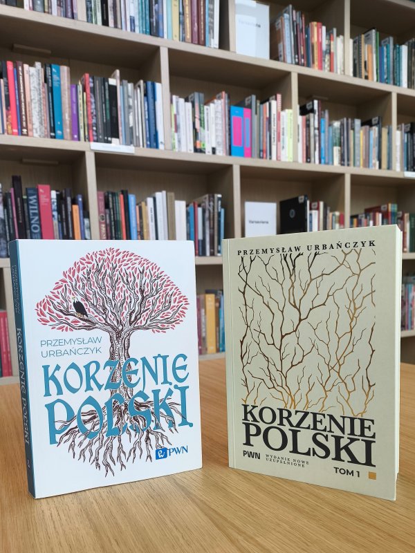 Dwie książki Przemysława Urbańczyka pt. „Korzenie Polski” stoją na drewnianym stole na tle regałów wypełnionych książkami. Po lewej stronie wersja z kolorową grafiką drzewa, po prawej tom 1 w jasnej okładce z grafiką nagich gałęzi.