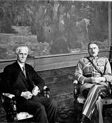 Na fotelach, na tle wielkiego obrazu siedzą Daszyński i Piłsudski. Daszyński jest w czarnym garniturze, a Piłsudski w mundurze wojskowym.
