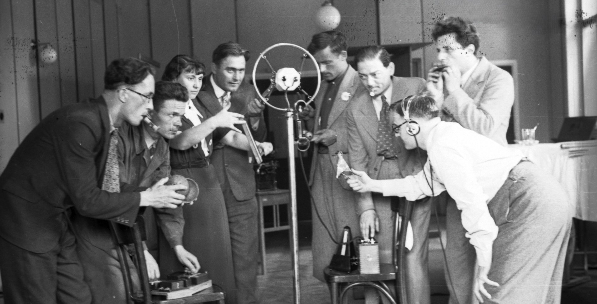 Czarno-białe zdjęcie przedstawia grupę ludzi w studiu radiowym skupionych wokół mikrofonu. Mężczyźni i kobieta w strojach z epoki imitują dźwięki za pomocą rekwizytów, a po prawej stronie realizator w słuchawkach kontroluje nagranie.