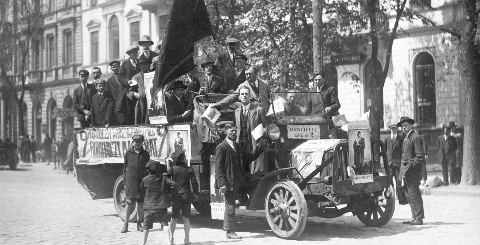Archiwalne czarno-białe zdjęcie z 1928 roku przedstawiające ciężarówkę agitacyjną Polskiej Partii Socjalistycznej na ulicy Warszawy. Pojazd jest obwieszony plakatami i czarnymi flagami, a wokół niego stoją mężczyźni roznoszący ulotki wyborcze.