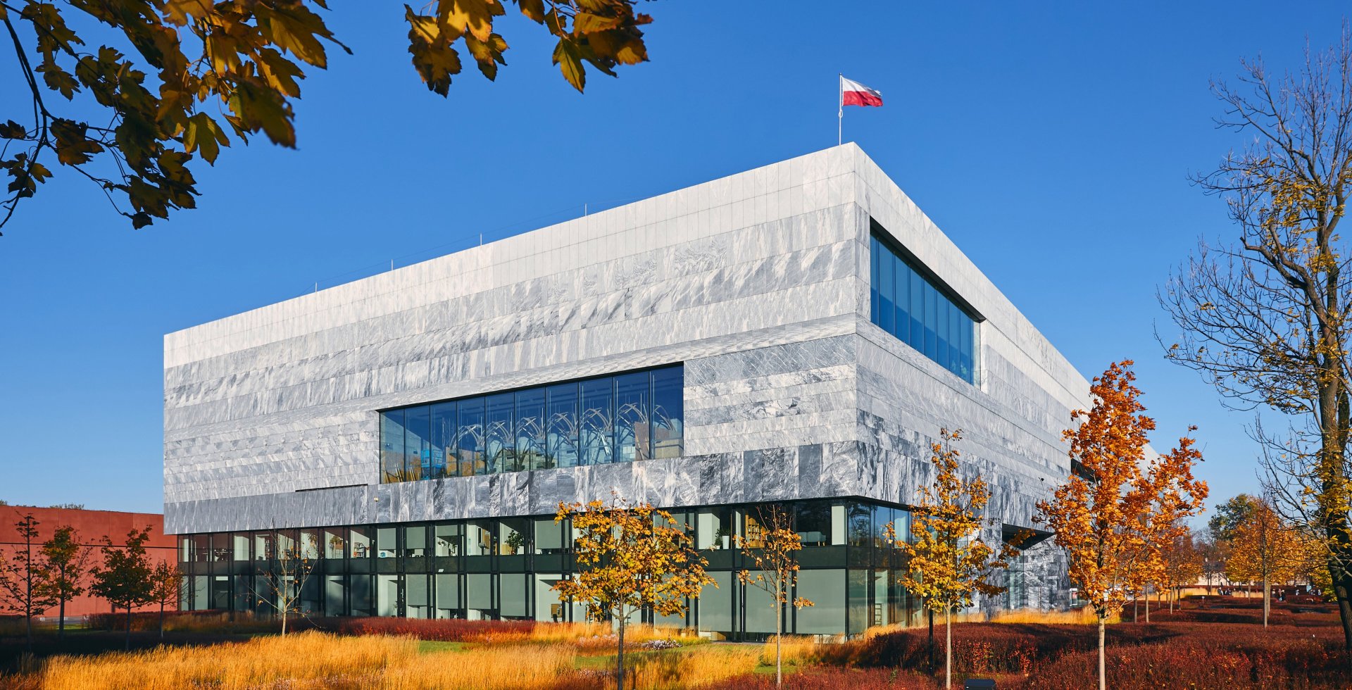Nowoczesny, monumentalny gmach Muzeum Historii Polski o jasnej, kamiennej fasadzie z geometrycznymi przeszkleniami. Na dachu powiewa polska flaga. Budynek otacza jesienna zieleń z żółto-pomarańczowymi drzewami pod bezchmurnym, niebieskim niebem.