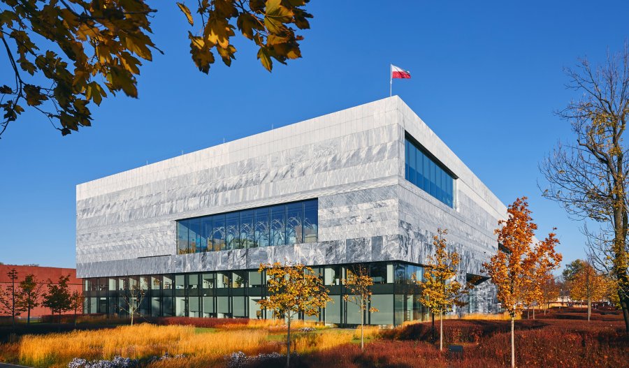 Nowoczesny, monumentalny gmach Muzeum Historii Polski o jasnej, kamiennej fasadzie z geometrycznymi przeszkleniami. Na dachu powiewa polska flaga. Budynek otacza jesienna zieleń z żółto-pomarańczowymi drzewami pod bezchmurnym, niebieskim niebem.