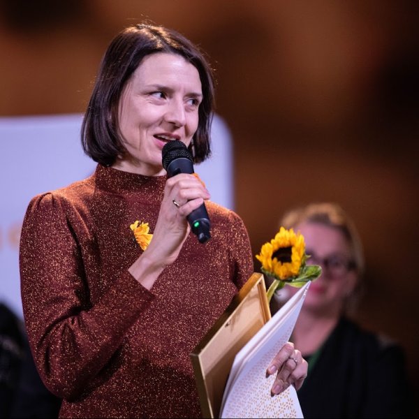 Uśmiechnięta kobieta o ciemnych włosach przemawia do mikrofonu, trzymając w rękach dyplom oraz żywy słonecznik. Ma na sobie połyskującą bluzkę w miedzianym kolorze. W rozmyte tle widoczne są sylwetki innych osób.