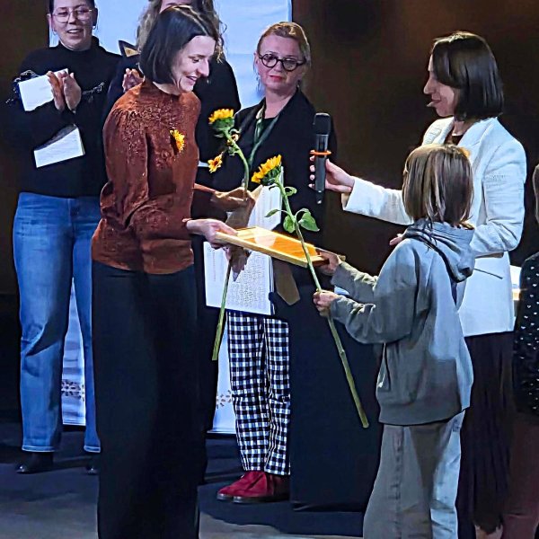 Uroczysta gala wręczenia nagród „Słoneczniki”. Dziecko wręcza dyplom oraz żywy słonecznik uśmiechniętej kobiecie na scenie. W tle stoją inne osoby, w tym klaszcząca kobieta, a powyżej widoczny jest fragment logo plebiscytu.